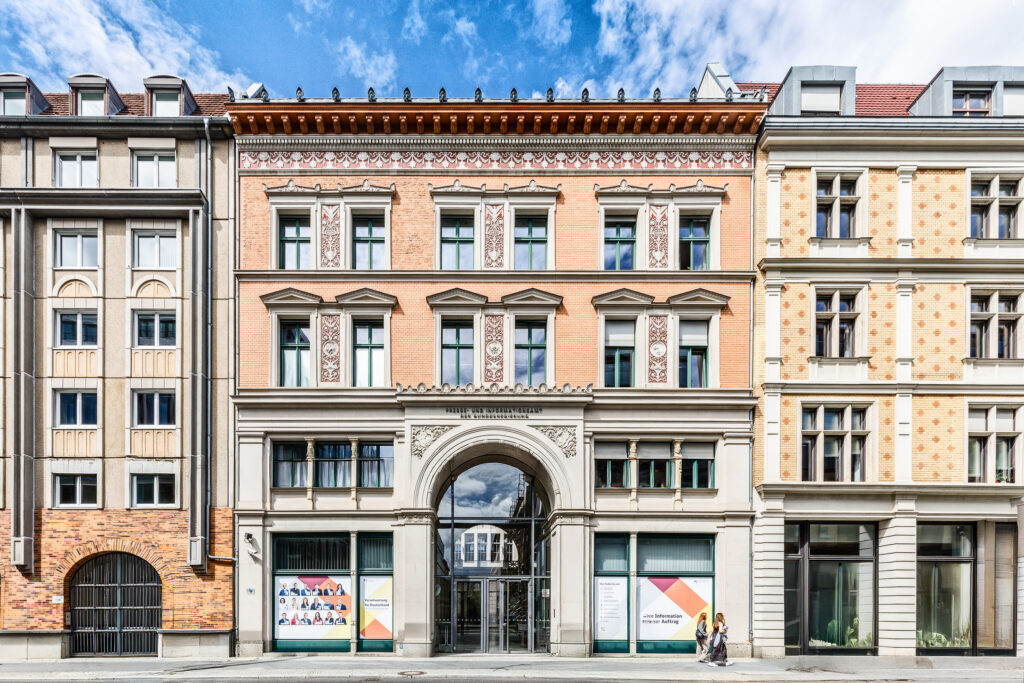 Historisches Gebäude mit farbigen Fassaden und dekorativen Elementen. Im Vordergrund gehen zwei Personen auf dem Bürgersteig vorbei.