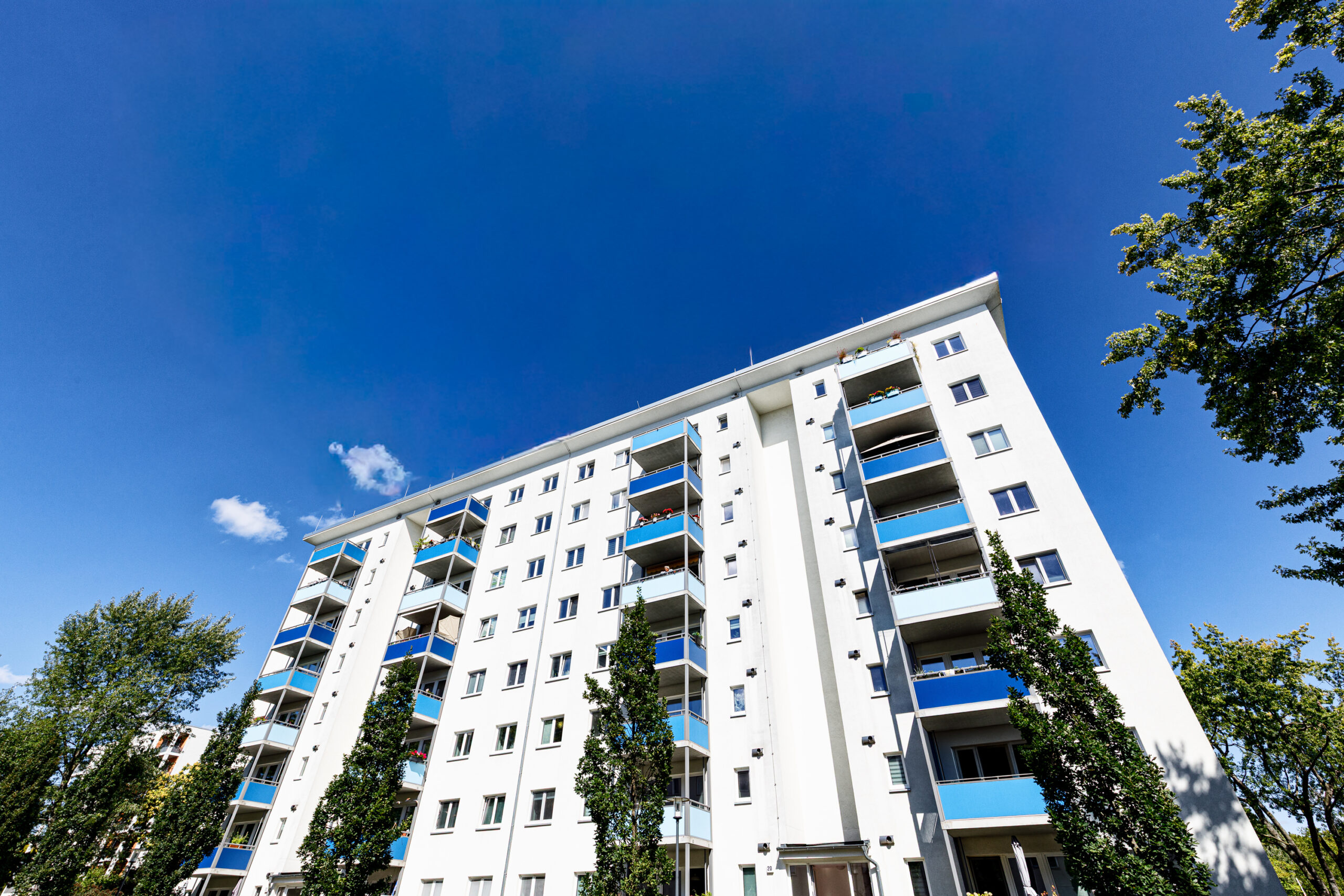 Moderne Wohnanlage mit fünf Stockwerken, weißen Wänden und blauen Balkonen, umgeben von Bäumen und klarem Himmel.