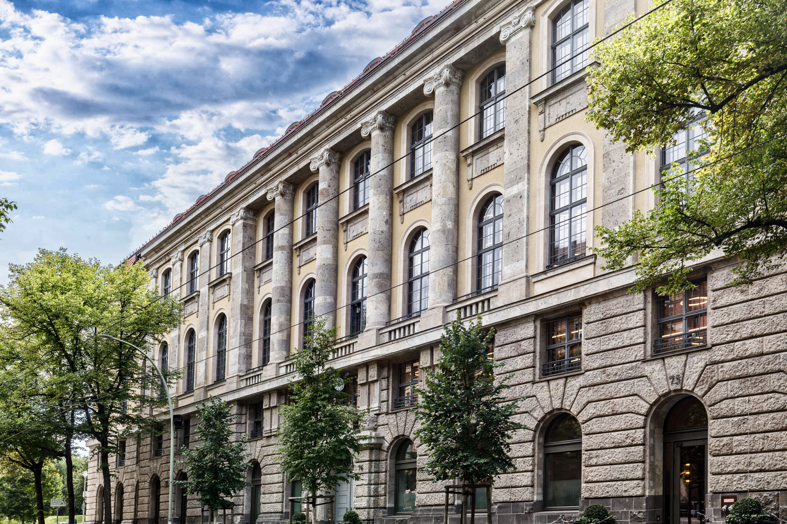 Historisches Gebäude mit Säulenfassade, großen Fenstern und grünen Bäumen im Vordergrund. Ideal für Architektur- und Stadtplanungskontexte.