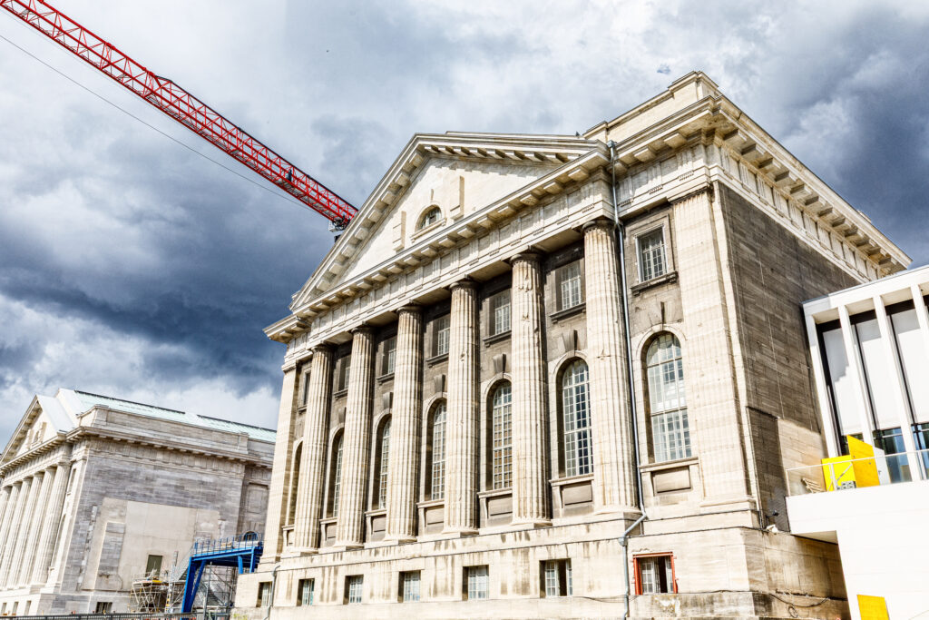 Historisches Gebäude mit Säulenfassade, im Hintergrund ein Kran und dunkle Wolken. Ideal für Architektur- oder Stadtentwicklungsprojekte.