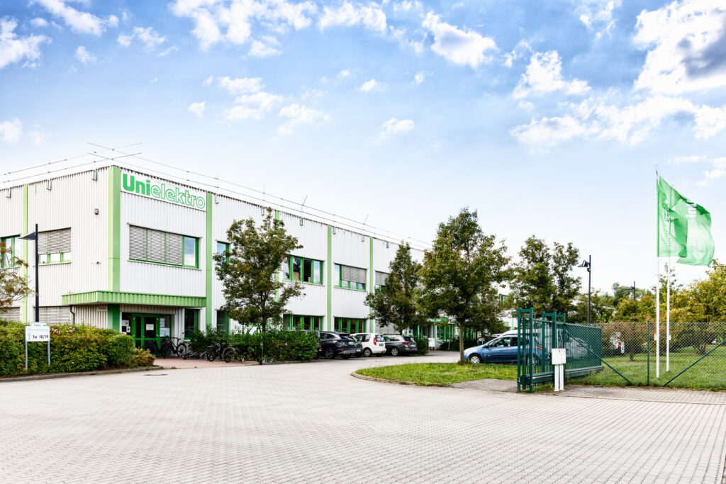 Fassade des UniElektro-Gebäudes mit grünen Akzenten, umgeben von Bäumen und einem Parkplatz.