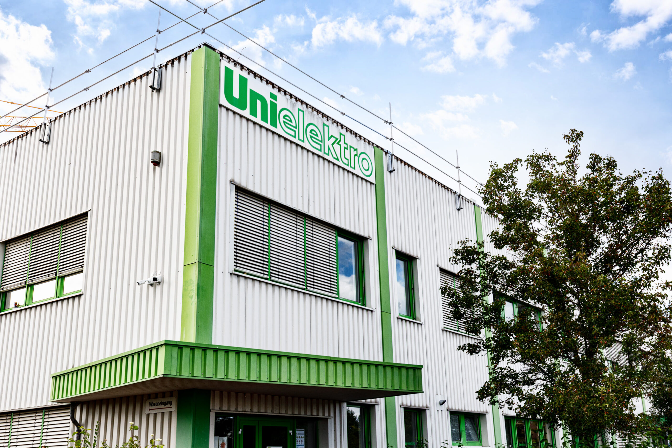 Fassade des UniElektro-Gebäudes mit grünen Akzenten und Fensterläden, umgeben von einem Baum.